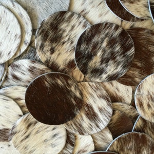 May include: A close-up of a pile of cowhide coasters in various shades of brown and white. The coasters are round and have a natural, rustic look.