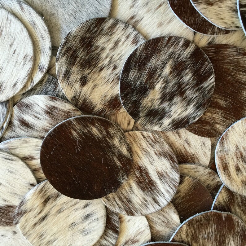 May include: A close-up of a pile of cowhide coasters in various shades of brown and white. The coasters are round and have a natural, rustic look.