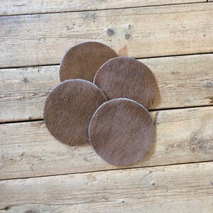 May include: Four brown cowhide coasters with a light brown color. The coasters are round and have a fuzzy texture.