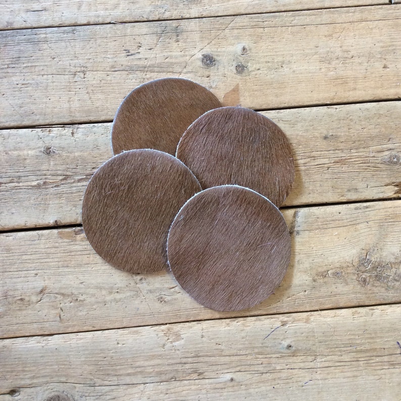 May include: Four brown cowhide coasters with a light brown color. The coasters are round and have a fuzzy texture.
