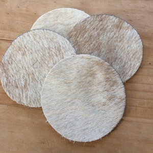 May include: Four round, white and brown cowhide coasters. The coasters are made of natural cowhide and have a soft, fuzzy texture.