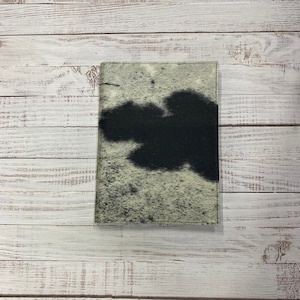 Puede incluir: Una cubierta de diario de piel de vaca negra y blanca con un diseño de salpicaduras negras.