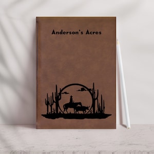 May include: Brown leather journal with the text "Anderson's Acres" embossed on the front cover. The journal features a silhouette design of a cowboy riding a horse with a steer in the background.