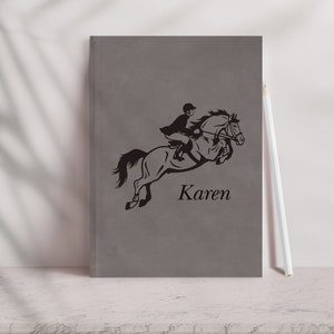 Pu&ograve; includere: Un quaderno grigio con una silhouette nera di una persona a cavallo che salta un ostacolo. Il nome "Karen" &egrave; stampato sotto l'immagine.