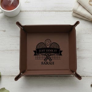May include: A brown leather tray with the text "JUST DINK IT" and the name "SARAH" in black. The tray has a pickleball design. A white mug with tea, a key, and folded napkins are also visible.
