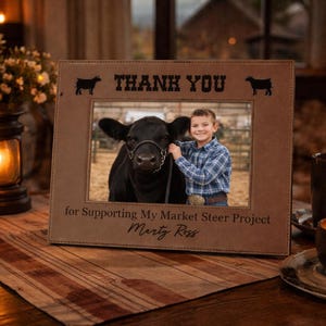 May include: A brown picture frame with the words "THANK YOU" and two cow silhouettes. The frame holds a photo of a boy and a black cow. The text "for Supporting My Market Steer Project Marty Ross" is below the photo.