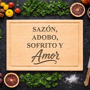 May include: A wooden cutting board with the words "SAZÓN, ADOBO, SOFRITO Y Amor" engraved on it. The board is surrounded by fresh ingredients like tomatoes, grapes, herbs, and a knife, suggesting a culinary theme.