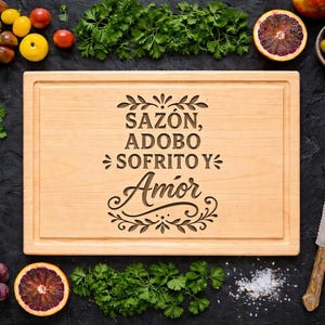 May include: A light wooden chopping board engraved with the words "Sazón, Adobo, Sofrito y Amor". The board is surrounded by fresh ingredients including tomatoes, grapes, herbs, and citrus fruits, suggesting a culinary theme.