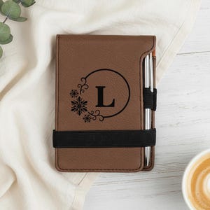 May include: Brown leatherette notebook cover with a black elastic band and pen holder. The cover features a black circular design with the letter "L" and snowflake accents. A silver pen is inserted in the holder.