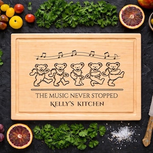 May include: A light-colored wooden cutting board with a carved design of dancing bears and musical notes. The text "THE MUSIC NEVER STOPPED KELLY'S KITCHEN" is below the bears. The board is surrounded by fresh produce and a knife.