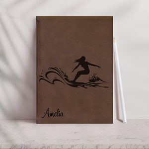 May include: Brown leather journal with a silhouette of a person surfing a wave and the name Amelia printed on the front.