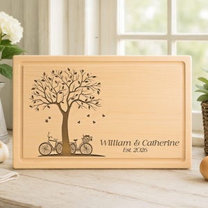 May include: A rectangular wooden cutting board with a tree and bicycle design. The tree is engraved with leaves and butterflies. The names "William & Catherine" and the year "Est. 2026" are also engraved.