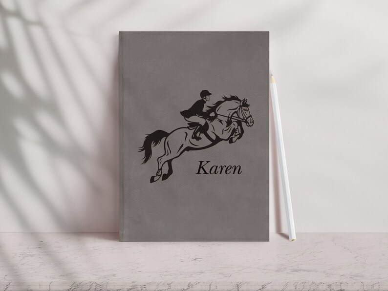 Pu&ograve; includere: Un quaderno grigio con una silhouette nera di una persona a cavallo che salta un ostacolo. Il nome "Karen" &egrave; stampato sotto l'immagine.