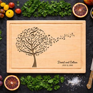 May include: A light-colored wooden cutting board with a tree design made of musical notes. The board has a rectangular shape with a recessed edge. The text "David and Colleen JULY 12, 2010" is engraved on the board. The board is surrounded by fresh produce.