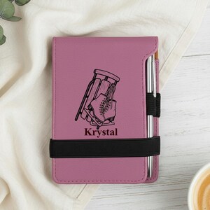 May include: A mauve notebook with a black elastic band and a silver pen. The notebook features a black graphic of ice skates and a sled, with the name "Krystal" below. The notebook is on a white surface.