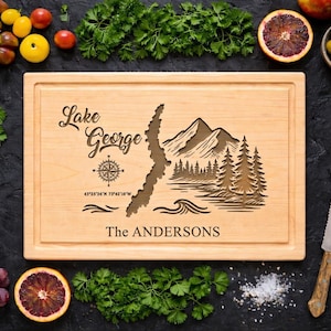 May include: A light-colored wooden cutting board with a carved map of Lake George, mountains, and trees. The text "Lake George" and "The Andersons" are engraved on the board. The board is surrounded by fresh produce.