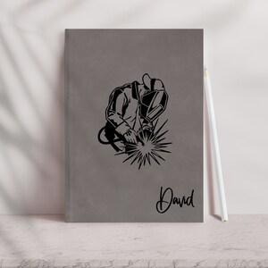 May include: Gray notebook with a black silhouette of a welder wearing a welding mask and holding a welding torch. The notebook has the name "David" written on the front.