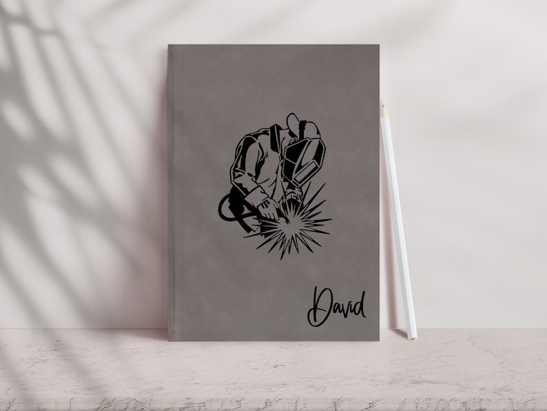 May include: Gray notebook with a black silhouette of a welder wearing a welding mask and holding a welding torch. The notebook has the name "David" written on the front.