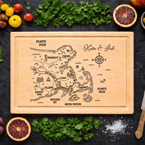 May include: A wooden cutting board with a map of Cape Cod and surrounding areas, engraved with the names "Katie & Josh". The board is surrounded by fresh produce, including tomatoes, grapes, and herbs. A knife and onion are also present.