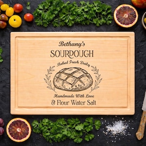 Peut inclure: Planche à découper en bois clair avec le texte "Bethany's SOURDOUGH" et une illustration de pain. Entourée de produits frais, dont des tomates, des raisins et des herbes, sur une surface sombre.