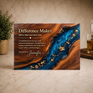 Peut inclure: Une plaque rectangulaire en bois avec un motif de grain naturel et une incrustation centrale en résine bleue. Le texte indique : Difference Maker [dif-er-uhns mey-ker] noun. A remarkable person who turns small actions into lasting impact. Also known as: Jennifer.