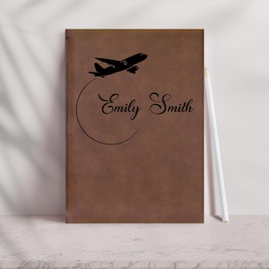 May include: Brown leather journal with a black airplane graphic and the name "Emily Smith" in elegant script. A white pencil rests beside the journal. The journal is a travel-themed item.