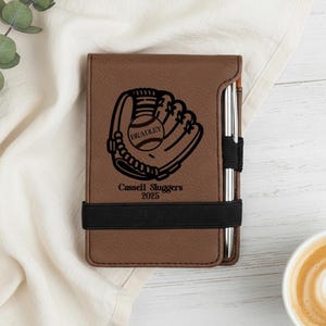 May include: Brown leather notebook with a baseball glove and ball design. The notebook has the name "Bradley" on the ball and the text "Cassell Sluggers 2025". A pen is held in place by a black elastic band.