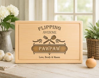 May include: A rectangular wooden cutting board with the words "FLIPPING AWESOME" and "PAWPAW" engraved on it. A fork and spoon are crossed above the word "PAWPAW". The text "Love, Brody & Mason" is below. The board is surrounded by a light-colored frame.