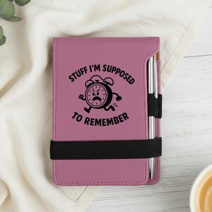 May include: A mauve notebook with the text "STUFF I'M SUPPOSED TO REMEMBER" and a running clock graphic. A black elastic band holds the notebook closed. A silver pen is inserted in the side.