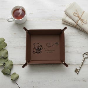 May include: A brown leather tray with a Winnie the Pooh design and the text "You will always bee my baby boy." A white mug with tea, a key, and napkins are also present.