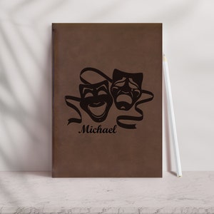 May include: Brown leather journal featuring a black design of two theatre masks and the name "Michael" engraved on the front.