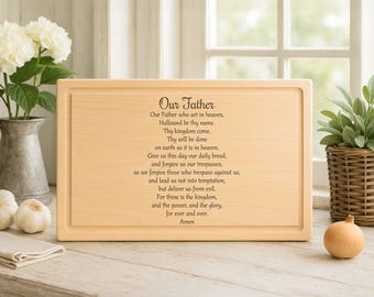 May include: A rectangular wooden cutting board with a carved border. The text of the Lord's Prayer is etched in black script on the board. The board is light brown and sits on a wooden surface.