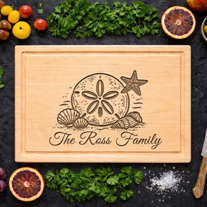 May include: A light-colored wooden cutting board with a carved beach-themed design, including a sand dollar, starfish, and seashells. The text "The Ross Family" is below the design. The board is surrounded by fresh produce.