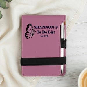 May include: A mauve notepad featuring the text "SHANNON'S To Do List" and a butterfly design. A black elastic band holds the notepad closed, and a silver pen is inserted in the side. The notepad is on a white surface.