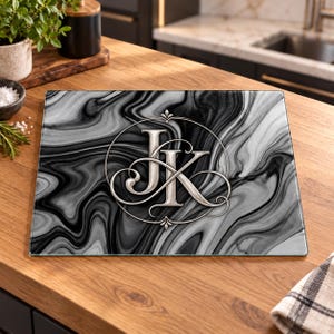 May include: A rectangular glass cutting board featuring a black and white marbled pattern. Centered on the board is a silver-toned circular emblem containing the stylized, interconnected capital letters J and K in a decorative script font.