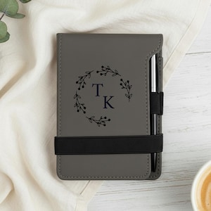 May include: Grey notepad with a black elastic band and a silver pen. The notepad features a floral wreath design with the initials "T K" in navy blue. The notepad is on a white surface.
