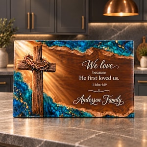 May include: A rectangular wooden wall plaque featuring a central cross with a crown of thorns. The design includes blue and gold resin accents and the text: We love because He first loved us. 1 John 4:19 Anderson Family, set against a wood grain background.
