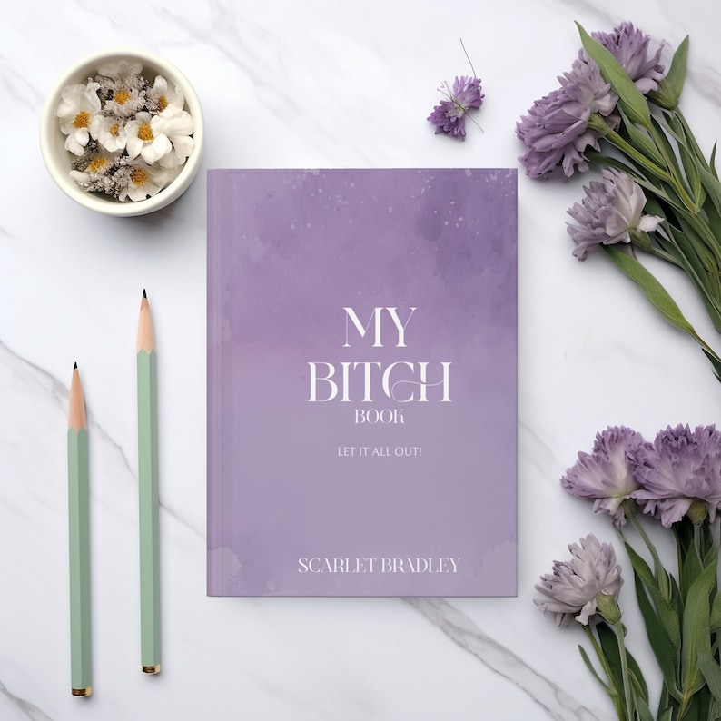 May include: A purple journal with the text "My Bitch Book Let It All Out!" and the author's name "Scarlet Bradley" on the cover. The journal is on a white marble surface with two green pencils and purple flowers.