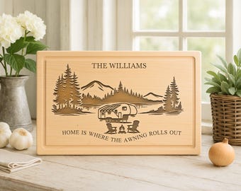 May include: A rectangular wooden cutting board with a carved camping scene. The design includes trees, mountains, a camper, and the text "Home is where the awning rolls out." The family name "The Williams" is above the scene.