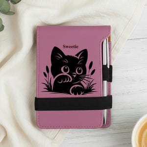 May include: A mauve-colored notebook with a black cat graphic and the word "Sweetie". The notebook has a black elastic band and a silver pen. The cat graphic is surrounded by grass.