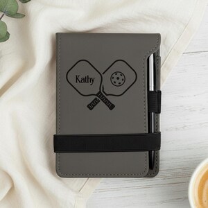 May include: Grey personalised pickleball notebook with a black elastic band and a pen holder. The notebook features a graphic of two pickleball paddles and a ball, with the name "Kathy" above the paddles.