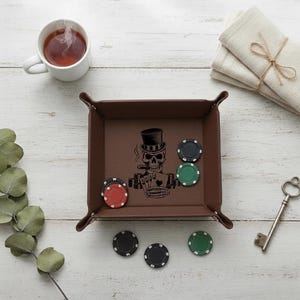 May include: A brown leather tray with a skull design, holding poker chips. The tray has a square shape with rounded corners. Poker chips in red, black, and green are scattered around the tray. A cup of tea and a stack of napkins are also present.
