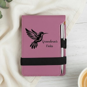 May include: A mauve notebook featuring a black hummingbird illustration and the words "Grandma's Notes." A black elastic band holds the notebook closed, with a silver pen inserted. The notebook rests on a white surface.