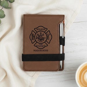 May include: A brown leatherette notepad with a black elastic band and a silver pen. The notepad features a black fire department emblem with the name "WILSON" above and "FIRE DEPARTMENT" and "MASON ROSS" below.