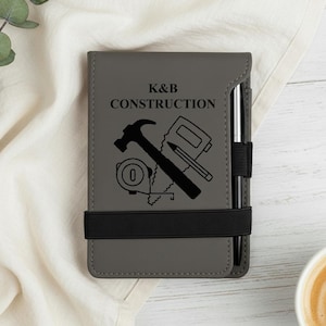 May include: Gray notepad with the text "K&B CONSTRUCTION" and construction tools. A black elastic band holds the notepad closed. A silver pen is inserted in the notepad's spine.