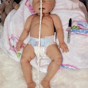 May include: A realistic newborn-style doll with light skin, closed eyes, and a neutral expression, wearing a disposable diaper. A white measuring tape placed vertically over the doll indicates a length of approximately 22 inches.