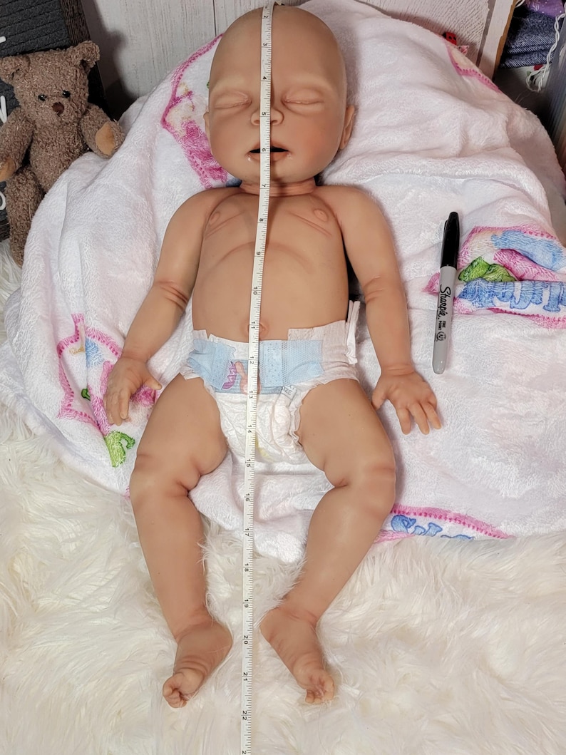 May include: A realistic newborn-style doll with light skin, closed eyes, and a neutral expression, wearing a disposable diaper. A white measuring tape placed vertically over the doll indicates a length of approximately 22 inches.