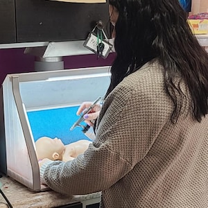 May include: A person wearing a beige textured cardigan uses a silver airbrush tool to paint a small, light-skinned doll head inside a portable, illuminated spray booth with a bright blue interior back panel and white side panels.