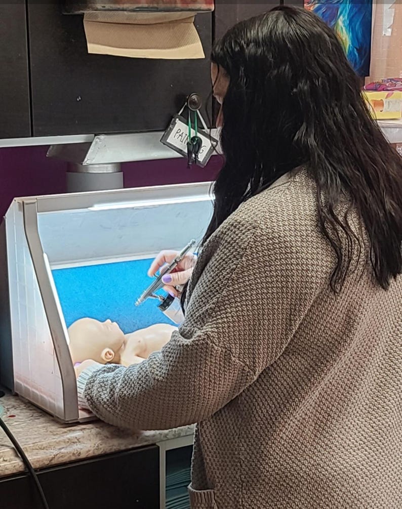 May include: A person wearing a beige textured cardigan uses a silver airbrush tool to paint a small, light-skinned doll head inside a portable, illuminated spray booth with a bright blue interior back panel and white side panels.