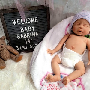 May include: A realistic newborn doll wearing a white diaper and a blue and white striped beanie, lying on a white blanket. Beside the doll is a brown plush teddy bear and a black letter board with the text WELCOME BABY SABRINA 14" 3 LBS 307.